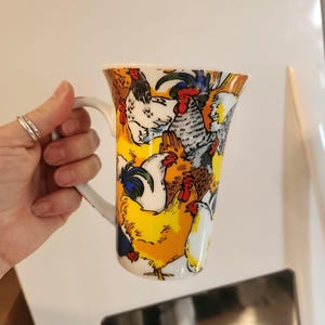 May include: A white ceramic mug with a colorful design of chickens and roosters. The mug has a tall, cylindrical shape and a handle.