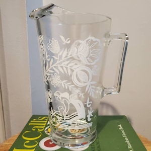 May include: A clear glass pitcher with a white floral design. The design features flowers, leaves, and a bird.