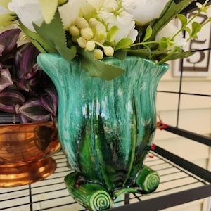 May include: A glossy, emerald green ceramic vase with a flared rim and a marbled finish. The base features three distinct, curled scroll-like feet. The vase is filled with a bouquet of white artificial flowers and green foliage.