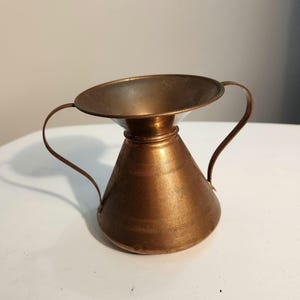 May include: A copper-colored, vintage-style pitcher with a wide, shallow top and a conical base. It features two curved handles and a decorative band around the middle. The pitcher is made of metal and has a polished finish.
