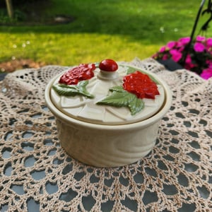May include: A cream-colored ceramic jar with a lid featuring red rose and green leaf accents. The lid has a small red knob. The jar has a textured, wood-like appearance. The jar is sitting on a beige lace doily.