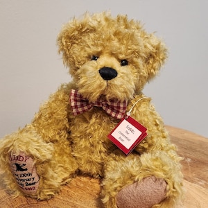 May include: A golden-brown teddy bear with a red and white checkered bow tie. The bear has a black nose and a tag that reads "Teddy, The Centennial Bear." The bear is sitting on a wooden surface.