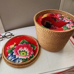 May include: A woven rattan wastebasket with a red floral fabric lining and lid. The lid features a large pink and white flower design with green leaves. The wastebasket is open, revealing the red floral interior.