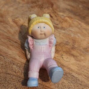 May include: A ceramic Cabbage Patch Kids doll figurine. The doll has blonde hair in pigtails, blue eyes, and is wearing a light blue shirt and pink overalls with blue shoes. The figurine is resting on a wooden surface.