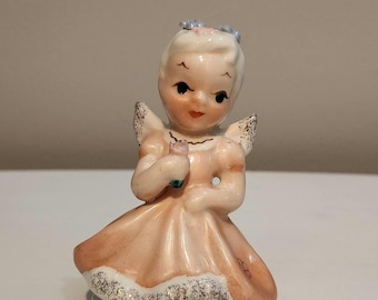 May include: A vintage porcelain figurine of a child angel in a peach-colored dress, holding a small flower. The figurine has delicate wings and a blue floral headpiece. The base reads "Tuesday's Child".