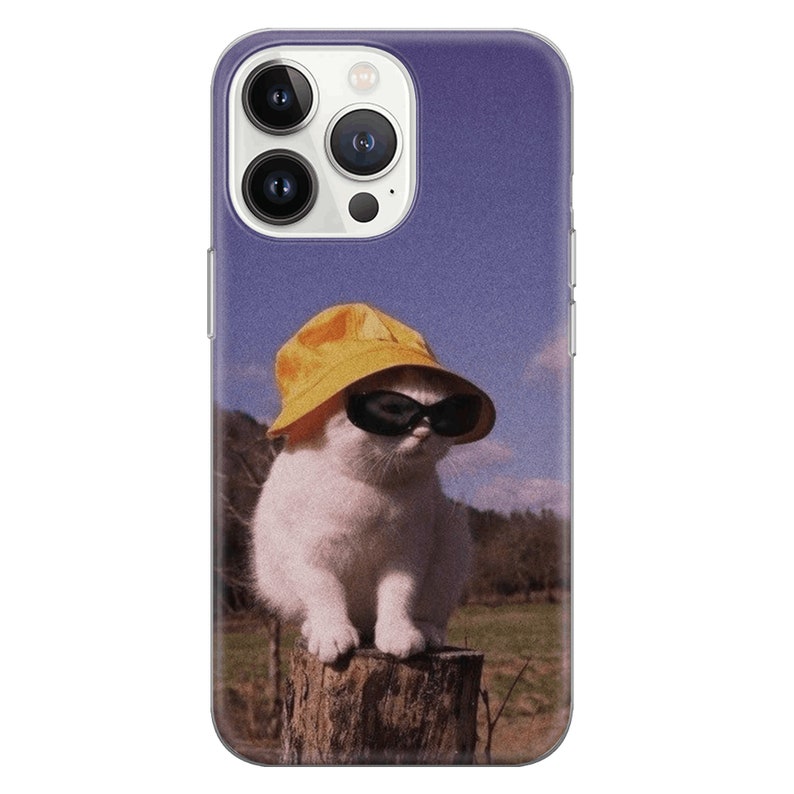 May include: A white cat wearing a yellow bucket hat and black sunglasses sits on a wooden stump. The cat is looking to the right. The background is a blurry image of a blue sky and green grass.