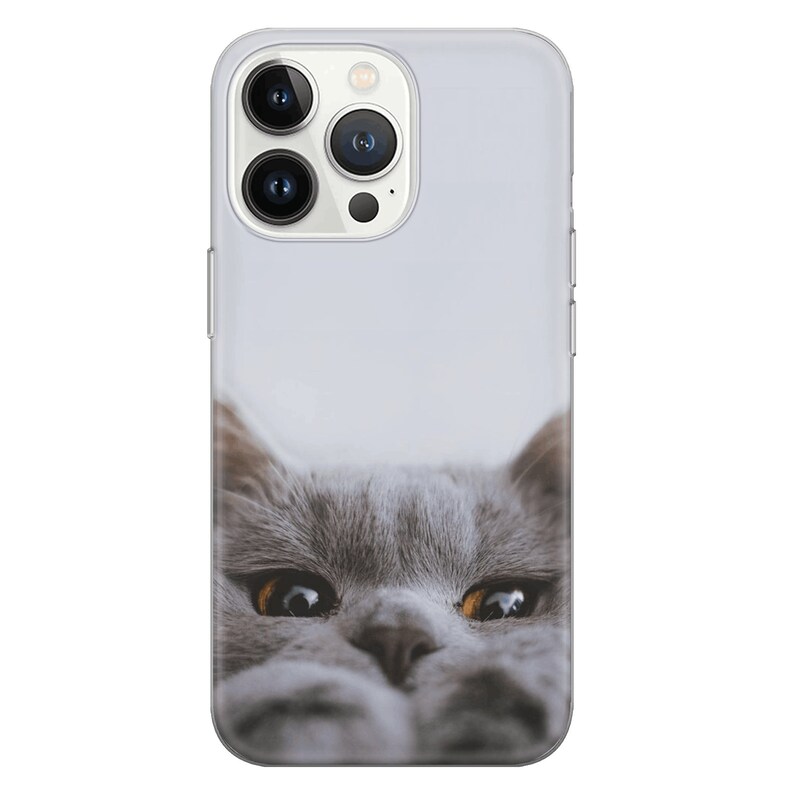 May include: A gray cat with yellow eyes peeks out from the bottom of a clear phone case. The case is designed for an iPhone with a camera bump on the back.