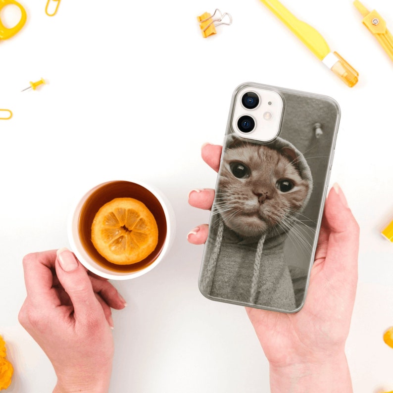 May include: A clear phone case with a photo of a cat wearing a gray hoodie. The cat has brown fur and big, round eyes.