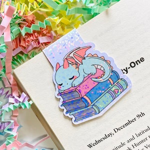 Peut inclure: Marque-page fantaisiste avec un dragon bleu endormi sur une pile de livres colorés. Le marque-page est inséré dans un livre ouvert avec le texte "ty-One". Le fond est orné de confettis colorés.