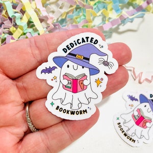 May include: A white ghost wearing a purple witch hat and holding a pink book. The ghost has a happy face and the text "Dedicated Bookworm" is written below the ghost.