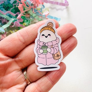 Peut inclure: Un personnage de dessin animé blanc portant un manteau d'hiver rose, un chapeau orange et tenant une tasse de café verte. Le personnage a un petit sac brun sur l'épaule.