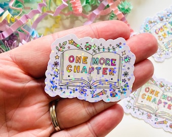 May include: A white sticker with a holographic finish features a colorful illustration of an open book with the text "ONE MORE CHAPTER" inside. The sticker is surrounded by a border of stars, flowers, and other whimsical designs.
