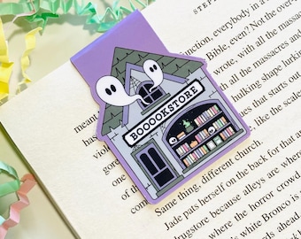 Op de afbeelding: Een paarse en witte bladwijzer met een cartoonillustratie van een spookwinkel met twee geesten. De winkel heeft een bord met de tekst "BOOOOKSTORE".