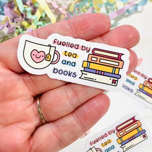 Peut inclure: Un autocollant blanc avec une tasse à thé rose avec un cœur et un sachet de thé, et une pile de livres colorés. Le texte sur l'autocollant est "Fuelled by tea and books".