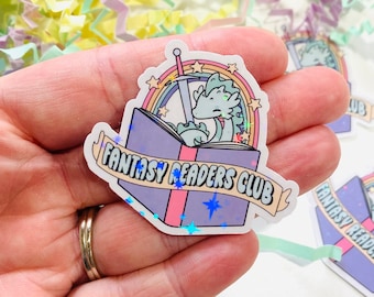 May include: A holographic sticker featuring a light blue dragon with a sword, sitting inside an open book with a rainbow and stars. The book is purple and pink with a white banner that reads "Fantasy Readers Club".