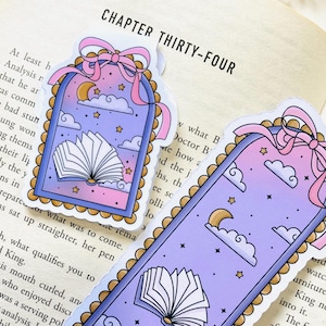 May include: Two whimsical bookmarks with a dreamy design. One is rectangular, the other is arched. Both feature an open book, clouds, stars, and a crescent moon against a purple and pink gradient. Each has a pink bow.
