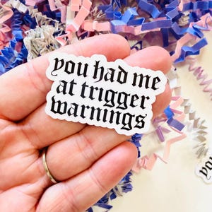 Peut inclure: Un autocollant blanc avec la phrase "you had me at trigger warnings" en police gothique. L'autocollant est tenu dans une main, entouré de papier déchiqueté bleu, rose et violet.