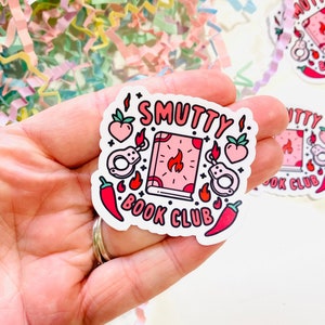 May include: A white sticker with the text "SMUTTY BOOK CLUB" in pink and black lettering. The sticker features a pink book with a flame, handcuffs, chili peppers, and peaches.