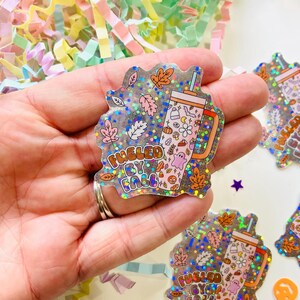 May include: A holographic sticker with a pink and white tumbler with the text "Fueled by Fall" and fall-themed illustrations. The sticker is on a hand.