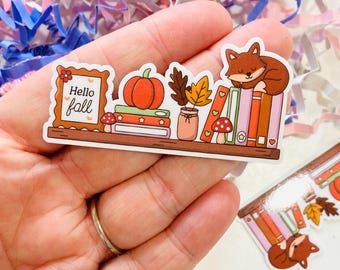 Puede incluir: Una pegatina con una estantería con elementos otoñales. Una calabaza, hojas y un zorro durmiendo están en la estantería. El texto "Hello Fall" está en un marco. La pegatina se sostiene en una mano.