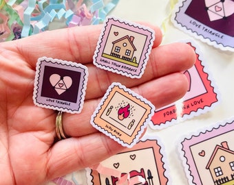 May include: A set of colorful stickers with various illustrations and text. The stickers are in the shape of postage stamps and feature designs such as a house, a heart, a fire, and a bed. The text on the stickers includes "Small Town Romance", "Love Triangle", "Slow Burn", "One Bed", and "Forbidden Love".
