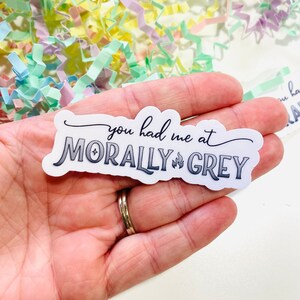 May include: A white sticker with the text "you had me at morally grey" in black lettering with a small flame icon between the words "morally" and "grey".