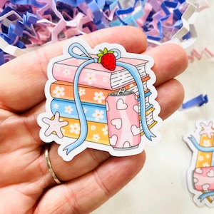 May include: A sticker featuring a stack of books with a strawberry on top, a pink soda can with heart shapes, and a starfish. The books are in shades of pink, orange, and blue, with a blue ribbon. The sticker is on a white background.