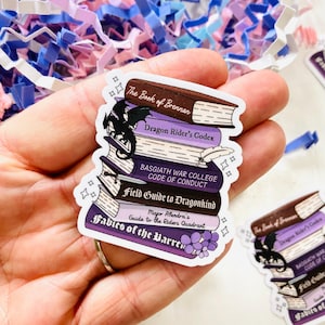 May include: A collection of fantasy book stickers featuring titles such as "The Book of Brennan" and "Dragon Rider's Codex." The stickers are predominantly purple, brown, and white, with a black dragon silhouette and floral details. The stickers are held in a hand.