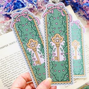 May include: Several decorative bookmarks with a key design. The bookmarks feature a green background with floral and key illustrations, framed by a scalloped edge. Each bookmark has a pink bow at the top.