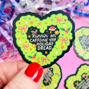 Può includere: Un adesivo a forma di cuore con un design a ghirlanda verde e la scritta "Runnin' on CAFFEINE and HOLIDAY DREAD." L'adesivo ha un bordo nero e presenta fiocchi, bacche e un cappello di Babbo Natale. Altri adesivi sono visibili sullo sfondo.