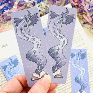 Op de afbeelding: Twee lavendelkleurige bladwijzers met een fantasiethema. Elke bladwijzer heeft een zwaard, vleugels en de tekst "The last page not yield until". Het ontwerp bevat een open boek en een sterrenspoor.