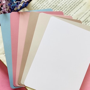 May include: A stack of blank, rectangular cards in various pastel colors, including blue, pink, and beige. The cards have rounded corners and are stacked on top of an open book. The image is taken from a slightly elevated angle.