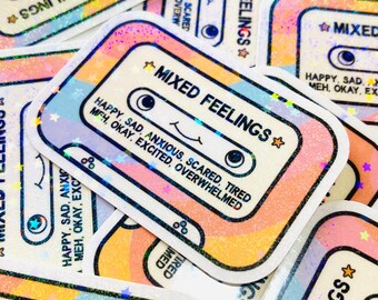May include: A close-up of several holographic stickers with the text "MIXED FEELINGS" and a cassette tape design. The stickers are rainbow colored and have a glittery finish. The text on the stickers reads "HAPPY, SAD, ANXIOUS, SCARED, TIRED, MEH, OKAY, EXCITED, OVERWHELMED."