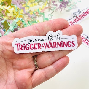 May include: White sticker with the text "give me all the TRIGGER WARNINGS" in black and pink lettering.