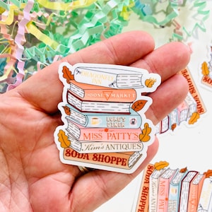 May include: A stack of colourful stickers featuring a stack of books with various business names, including "Dragonfly Inn", "Doose's Market", "Luke's Diner", "Miss Patty's", "Kim's Antiques", and "Soda Shoppe".