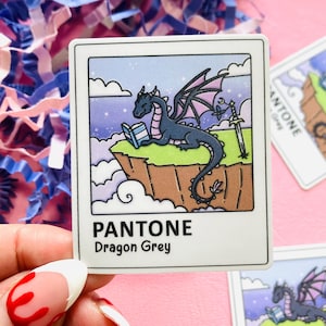 May include: A square sticker with a cartoon dragon reading a book on a cliff, with a sword. The sticker has a white border and the text "PANTONE Dragon Grey". The background is a mix of pink, blue, and white.