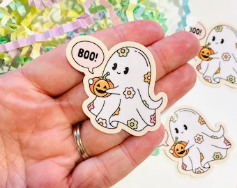 Pode incluir: Um adesivo de fantasma branco com um padrão floral, segurando uma pequena abóbora laranja e um balão de fala que diz "BOO!"