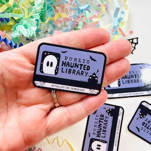May include: A purple and black sticker with the text "Public Haunted Library" and a ghost illustration. The sticker has the text "Afterlife Membership" at the bottom.
