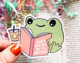 May include: A green frog wearing a pink shirt and holding a book that says "Don't Bug Me Or I'll Eat You". The frog is holding a cup of coffee in its other hand.