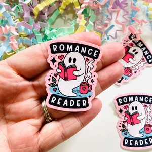 May include: A pink sticker featuring a cartoon ghost reading a book, with the text "ROMANCE READER". The ghost holds a red book and a mug with a heart. The sticker has a pink border and is decorated with hearts and stars.