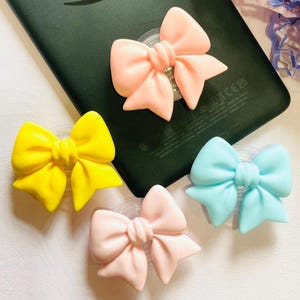 May include: A selection of bow-shaped phone grips in yellow, light pink, baby blue, and a darker pink. The bows are attached to clear, circular bases, designed to stick to the back of a phone or tablet. A black tablet is in the background.