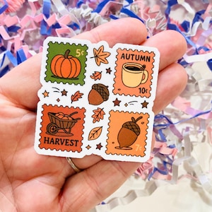 May include: A square sticker with four postage stamp designs, each with an autumnal theme. The stamps show a pumpkin, a coffee cup with the word "AUTUMN", a wheelbarrow with "HARVEST", and an acorn. The sticker has a white border.