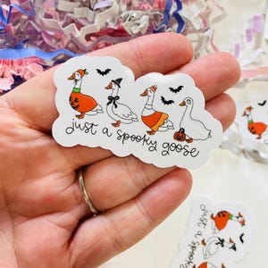 May include: A white sticker with the text "just a spooky goose" and illustrations of geese in Halloween costumes. The geese are wearing a pumpkin, a witch hat, and an orange skirt. Black bats are also included in the design.
