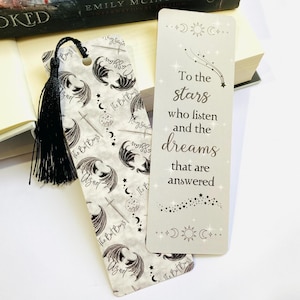 May include: A grey and white bookmark with a black tassel. The bookmark features a repeating pattern of black bat wings and a quote that reads "To the stars who listen and the dreams that are answered".