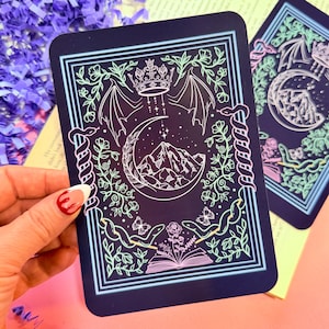 May include: A dark navy card with a detailed illustration featuring a crescent moon, mountains, a crown, and wings. The design is framed with a light blue border and floral elements. The card has a mystical theme.