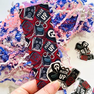 May include: A bookmark with a red background and a repeating pattern of gas masks, handcuffs, and the text "Mask Up". There are also three stickers with the same design.