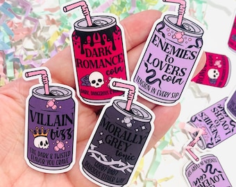 Puede incluir: Cinco pegatinas de lata de colores con diferentes diseños y textos. Las pegatinas presentan una calavera, una serpiente, una corona y alambre de púas. El texto en las pegatinas incluye "Dark Romance", "Enemies to Lovers", "Villain Fizz", "Morally Grey" y "Good Bad Delicious Either Way".