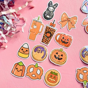 May include: A selection of Halloween stickers on a pink surface. The stickers feature a jack-o'-lantern, a candy corn, a ghost cookie, a pretzel, a bow, a skeleton bunny, and a coffee cup. The stickers are in orange, black, and white.