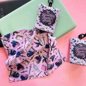 May include: A light pink microfiber cloth and two matching pouches with the text "Romantasy Readers Society". The cloth features a fantasy-themed pattern with swords, books, and dragons. The pouches have a floral design.