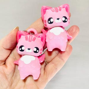 Può includere: Due statuette di gatto rosa con grandi occhi neri e segni bianchi sul petto. Le statuette di gatto hanno un portachiavi attaccato a una di esse. I gatti hanno orecchie rosa e strisce sulla testa. Le statuette sono tenute in una mano.